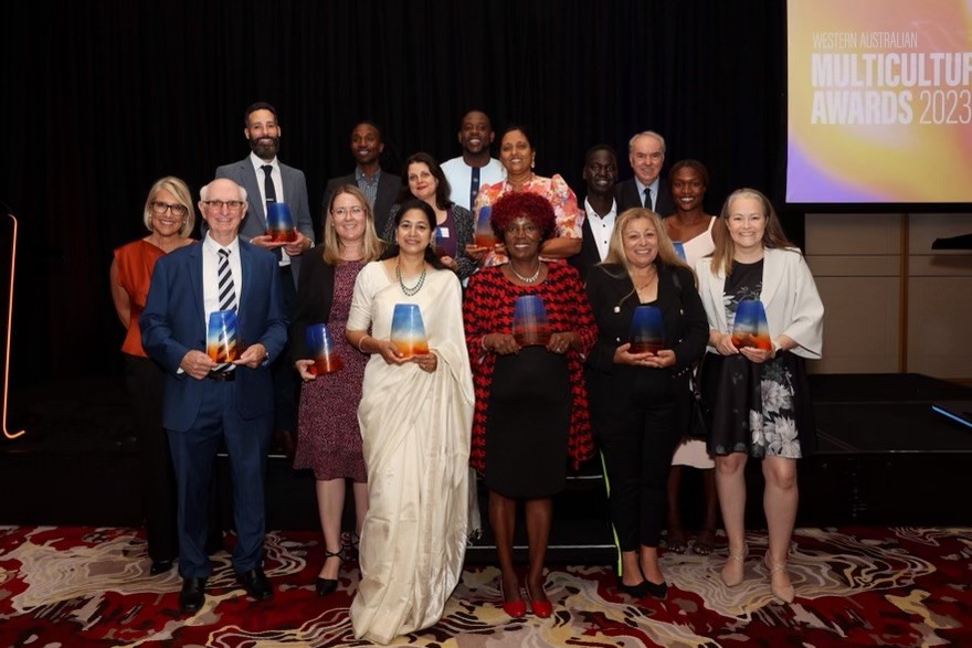 15 people standing on a stage smiling and holding glass trophies. Six are men and nine are women.  They are in front of a screen that says Multicultural Awards